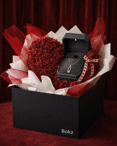 Black jewelry box with a necklace inside, surrounded by red flowers in a heart shape on a red background.